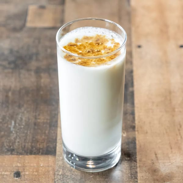A tall glass of white lassi with a golden-brown spice topping on a wooden table.