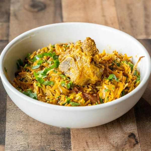 A white bowl filled with yellow biryani rice, topped with a piece of meat and fresh cilantro.