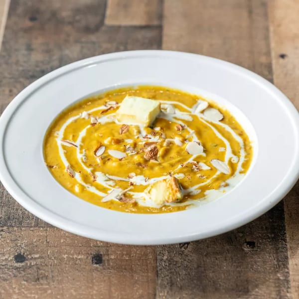 A bowl of creamy orange soup with white swirls, topped with croutons, almonds, and nuts.