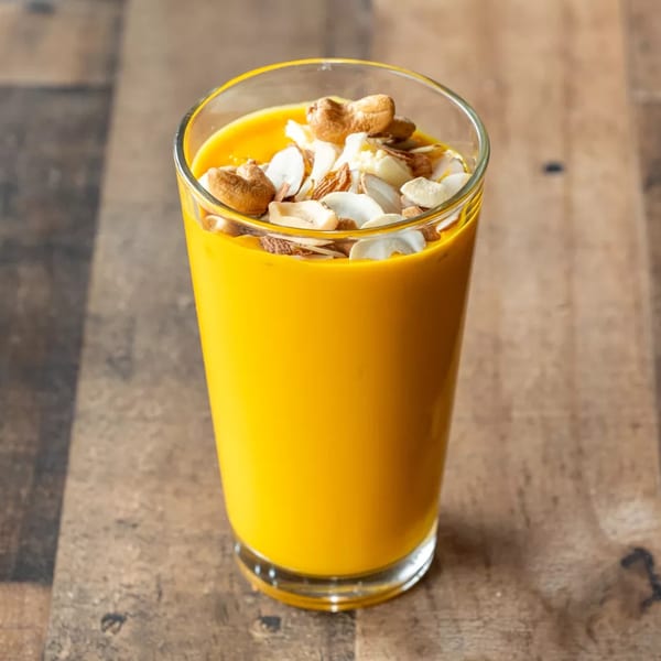 A vibrant orange mango lassi in a glass, topped with cashews and sliced almonds.