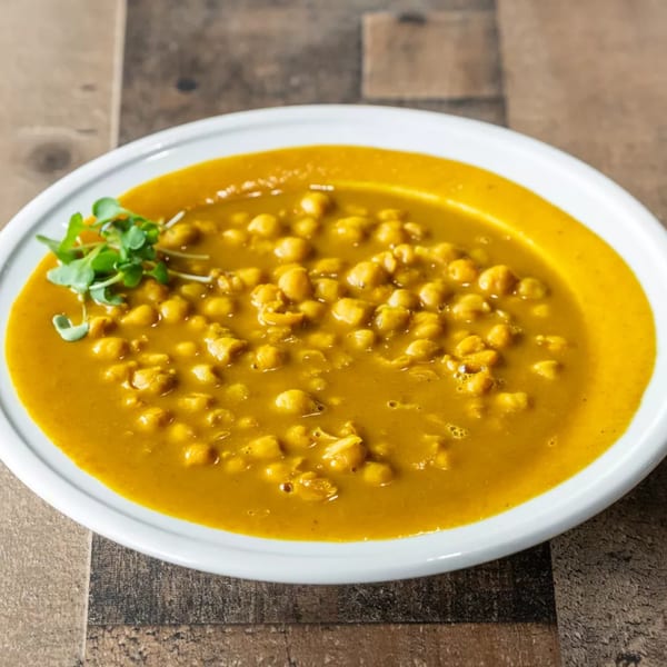 A white bowl filled with yellow chickpea curry, garnished with fresh green sprouts on a wooden table.