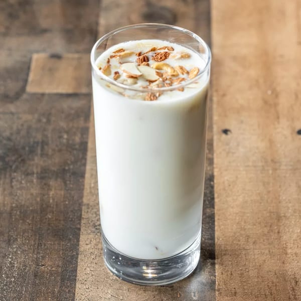 A tall glass of white lassi topped with sliced almonds and spices on a wooden table.
