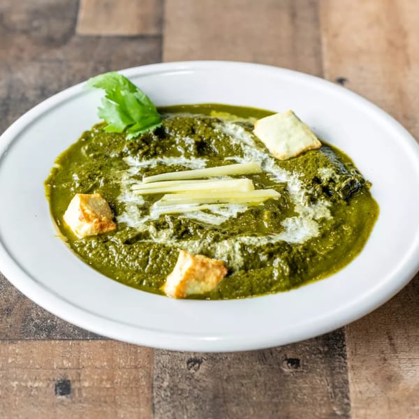 A white bowl of vibrant green palak paneer, garnished with cream, ginger strips, and cilantro.
